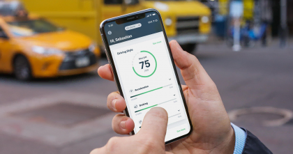 Un uomo tiene in mano uno smartphone che visualizza un'applicazione con un punteggio di guida (75/100) e metriche sullo stile di guida, come l'accelerazione e la frenata, evidenziando un comportamento di guida sicuro. Sullo sfondo è visibile una strada cittadina con un taxi e un veicolo giallo.