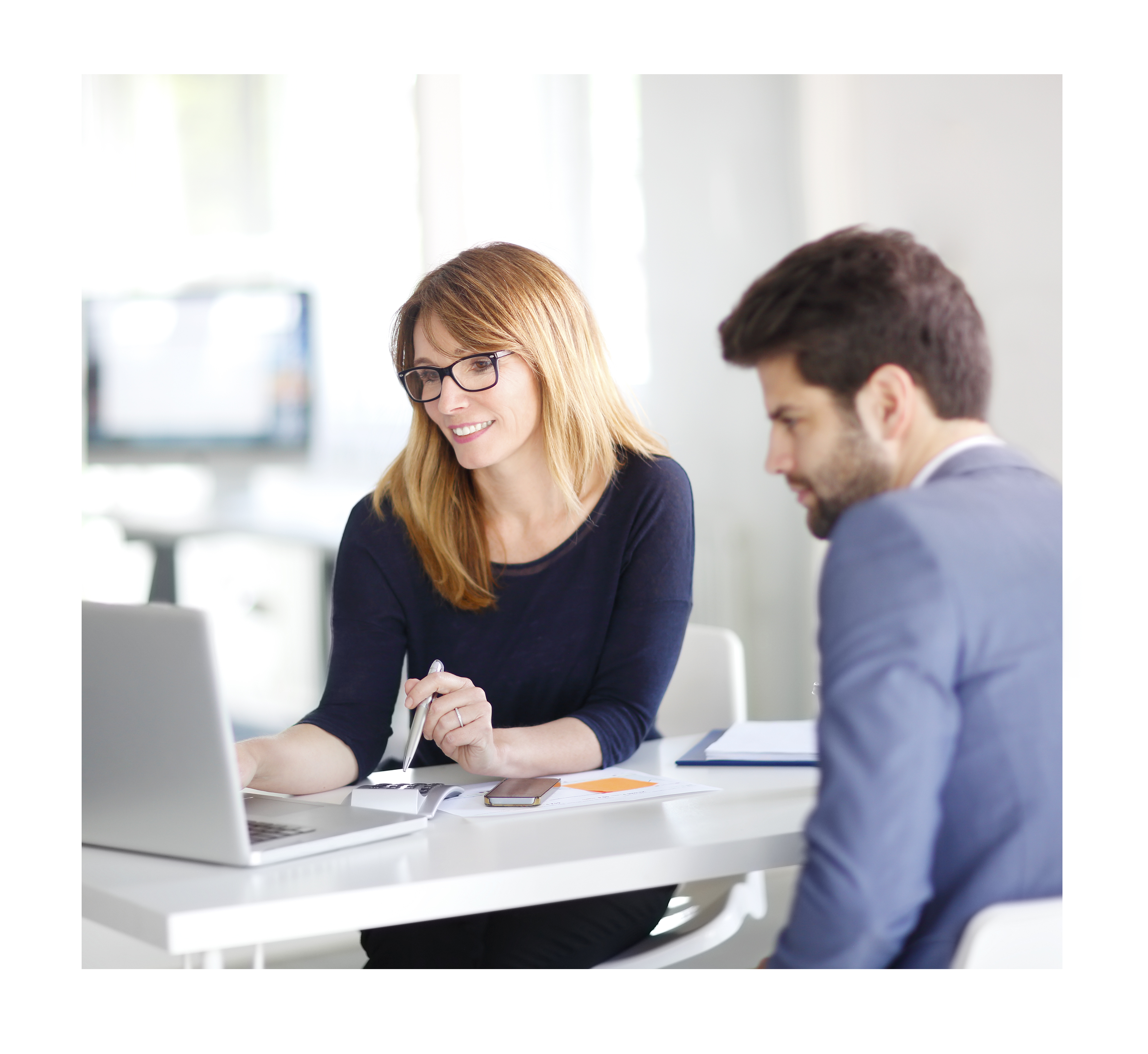 A professional woman wearing glasses smiles while presenting data on a laptop to a colleague seated next to her. The setting is a bright and modern office environment, emphasizing collaboration and effective communication