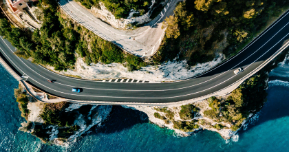 Aerial view of a scenic coastal road with sharp curves, surrounded by cliffs, lush greenery, and crystal-clear sea